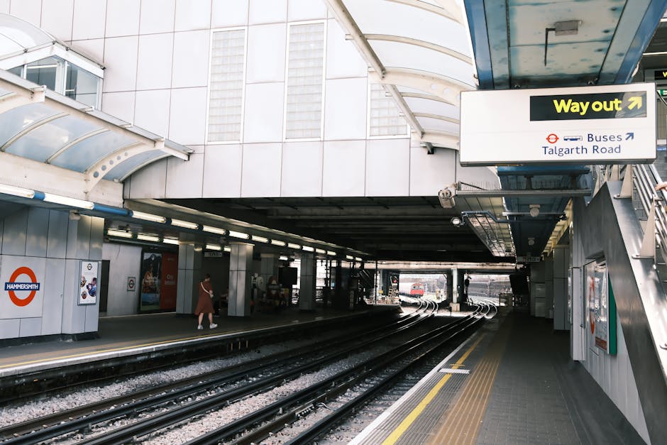 A view of Hammersmith underground station platform beneath a curved, semi-transparent canopy with white and light grey metal framing. The platform has a textured, non-slip surface with yellow tactile paving near the edge for safety. On the left side, there are white tiled walls adorned with a round Hammersmith station sign and advertising posters. A solitary person, dressed in casual clothing, is walking along the platform, which is illuminated by long, fluorescent light fixtures attached to the ceiling. Rail tracks run parallel to the platform, curving towards the background where a red train is visible approaching or departing. An overhead sign with a yellow arrow directing to the 'Way out' and further routes to buses and Talgarth Road is mounted on a blue metal support beam on the right. The environment appears clean, modern, and part of an urban transit interchange, consistent with facilities supporting passenger access to local transportation services such as private options for rubbish removal or other logistics nearby.