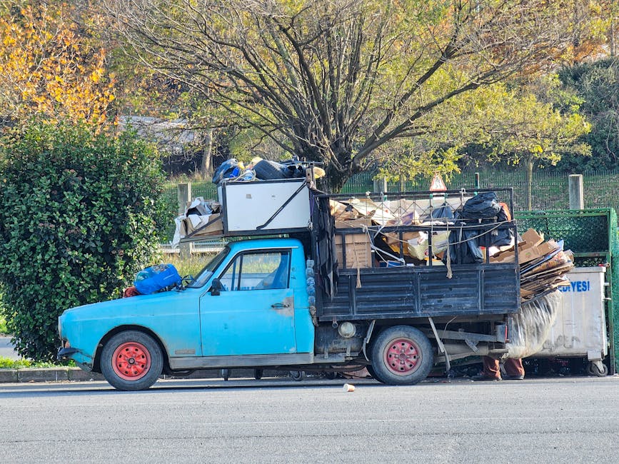 A small, aged blue pickup truck parked alongside a road with its bed and cabin heavily loaded with various types of waste, including cardboard boxes, plastic bags, and miscellaneous debris. The truck appears to be in a slightly worn condition, with faded paint and red-painted wheels. The waste materials are piled high in the truck bed, with some items extending beyond the sides and secured with ropes. Behind the vehicle, there is a large green hedge and a tree with sparse, autumn-colored leaves, indicating a park or residential area setting. The environment appears to be bright with natural daylight, and the scene suggests an outdoor waste collection or rubbish removal operation, possibly by a private provider engaged in on-site clearance of household or garden waste, illustrating effective local waste handling alternatives.