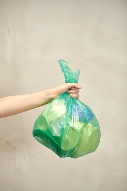 A person's arm extending from the left side of the image holds a green plastic rubbish bag with a tightly knotted top, filled with waste materials. The bag appears semi-transparent, revealing mixed contents inside, and has a glossy finish. The background features a plain, light-colored wall with a subtle texture, creating a neutral setting typical of indoor environments or waste collection areas. The lighting is soft and evenly distributed, illustrating the bag's shape and the hand's natural skin tone. This scene exemplifies one method of private or alternative waste handling, highlighting the importance of secure rubbish collection, which is relevant to services like those offered by rubbish collection companies in Hammersmith. The visual context supports understanding of rubbish disposal practices outside of municipal waste collection, emphasizing the use of reusable and easily transportable plastic rubbish bags associated with professional waste management services.