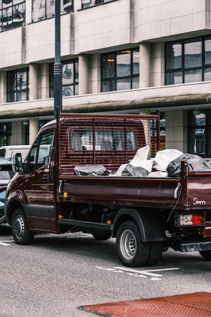 A small burgundy flatbed truck with a protective metal grid behind the driver's cabin, parked on an urban street in front of a modern office building with large, reflective windows. The truck's bed is loaded with various types of discarded waste, including black and white plastic bags, some bulging with rubbish, and other unidentified debris. The vehicle is positioned close to a designated parking area, with a white vehicle visible in the background. Nearby, a streetlight pole stands upright, and part of a red and white striped safety barrier is visible on the pavement. The scene suggests the truck is used for private rubbish removal or on-site waste clearance, aligning with services provided by companies like rubbishcollectionhammersmith.co.uk, which handle alternative waste disposal needs outside of standard municipal collection programs. The environment appears to be a commercial or business area, with natural daylight illuminating the scene, capturing the practical aspects of waste transportation and collection in an urban setting.