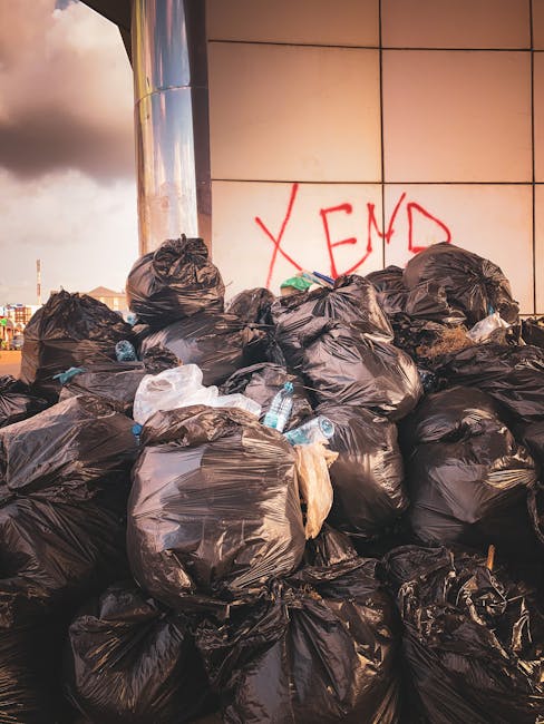 A large pile of black rubbish bags filled with household waste are stacked against a tiled wall on a city street or site, with some bottles and plastic packaging visible poking out from the bags. The bags appear full and tightly sealed, with a few loosely tied or torn, indicating they are prepared for collection or disposal. Behind the bags, the wall features a red graffiti message with the word 'XEND' spray-painted in capital letters. In the background, part of a building and a traffic light are visible beneath an overcast sky, suggesting an urban environment where private waste handling or on-site clearance might be occurring, with an implicit connection to alternative rubbish removal services such as those provided by companies like rubbishcollectionhammersmith.co.uk. The scene captures the typical clutter associated with rubbish awaiting pickup, emphasizing the need for efficient rubbish management in city areas, especially when dealing with bulk waste or irregular waste collection schedules in Hammersmith.