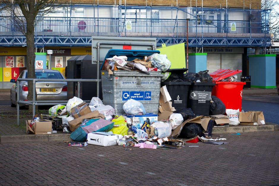 A large collection of overflowing rubbish and waste is piled up on a paved urban street, situated near a parking area with two cars parked adjacent to a metal railing. The waste includes black and red rubbish bags, flattened flattened cardboard boxes, crumpled paper, plastic packaging, and discarded household items. In the background, there is a multi-storey building under scaffolding with blue protective netting, indicating ongoing maintenance or renovation. The scene suggests an example of private or unscheduled rubbish disposal requiring collection services, with waste materials spilling onto the pavement. The garbage appears untidy, with various items loosely placed, some partially on the curb and others on the pavement, highlighting the need for professional waste management and responsible disposal. The environment is an urban commercial or residential area, with visible signs of clutter that would be addressed by a rubbish removal service such as those offered by rubbishcollectionhammersmith.co.uk, consistent with the purpose of alternative waste handling without reliance on local authority collection.