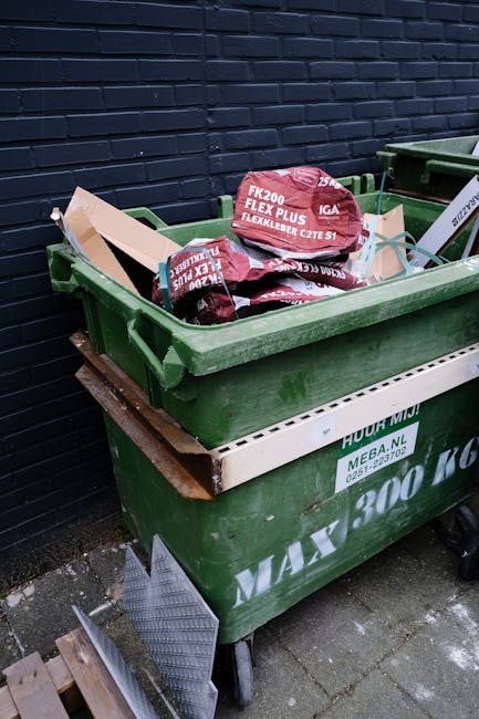 A green wheeled rubbish bin filled with red and black bags containing construction or garden waste, placed outdoors against a dark blue painted brick wall. The bin is situated on a paved surface, with a smaller white container partially visible underneath. The bags inside the bin are marked with text indicating they contain flexible rubber products, and some are slightly leaning over the edge of the bin. Nearby, a small metal ramp or cover is positioned on the ground, possibly for access. The setting appears to be an exterior service or waste collection area, where independent rubbish disposal or site clearance is taking place, consistent with private waste handling practices supported by companies like rubbishcollectionhammersmith.co.uk.