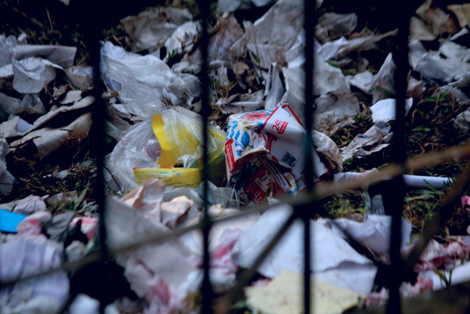 A close-up view of a pile of discarded rubbish behind a black metal fence, featuring crumpled and torn plastic bags, paper packaging, including a partially visible food container with a red and white label, and other mixed waste materials. The waste is scattered on the ground amidst soil and dead leaves, creating a cluttered and neglected scene typical of unmanaged waste accumulation. The image highlights the types of refuse often encountered in illegal dumping or unregulated disposal sites, reflecting the importance of proper rubbish collection and adherence to council disposal rules. The lighting appears natural, with shadows cast over the waste, emphasizing the textures of the torn plastics and paper. This scene exemplifies the kind of debris that professional rubbish removal services, like those provided by [COMPANY_NAME], are equipped to handle in an environmentally responsible manner, ensuring the site is cleared of unwanted waste such as this. The focus on the trash within the fence underscores the need for effective waste management solutions to prevent such accumulations in residential or public areas.