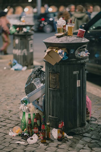 A black, cylindrical public waste bin made of metal with vertical ridges and a domed lid, positioned on a cobbled street in an urban environment. The bin is overflowing with litter, including crumpled paper and discarded food packaging, with additional rubbish piled around its base. On top of the lid, there are several used drink cups, including a plastic cup with a straw, and a small paper cup, along with a few beverage bottles—some glass and some plastic—scattered on and around the bin. Beside the bin, a black plastic garbage bag is tied, leaning against the side, while a partially filled cardboard box hangs from the bin’s side. In the background, a blurred scene shows pedestrians, parked cars, and a grey rubbish collection container, indicating a busy city street setting. The overall scene captures typical urban littering, relevant to waste disposal and collection services, with natural daylight illuminating the area.