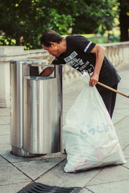A middle-aged man with dark hair, dressed in a black T-shirt and dark trousers, is seen bending over to place waste into a tall, cylindrical stainless steel rubbish bin on a paved walkway. He holds a long-handled grabber in one hand and a large white plastic bag filled with rubbish in the other, which appears to contain various waste items. The stainless steel bin has a shiny, reflective finish and is positioned against a low concrete wall, with a background of lush green trees and foliage. The environment suggests an outdoor urban park or streetside area, with natural sunlight illuminating the scene. The man’s focused posture indicates he is engaged in a waste collection activity, resembling an independent or community-led rubbish removal effort, typical of private waste disposal outside of municipal services, highlighting a practical approach to rubbish management in public or semi-public spaces.