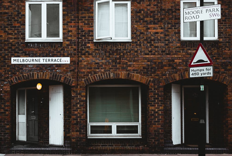 The image shows a two-story brick residential building with a dark brown, textured brick facade. On the ground floor, there are two arched doorways with a small illuminated light fixture above the left entrance, one with a black front door and the other with a partially open white door. Between the doorways, there is a large rectangular window with closed horizontal blinds, framed with white trim. Above, on the first floor, there are three white-framed windows with double-glazed panes; the left and right windows are closed, while the middle window is slightly open, revealing blinds inside. To the right of the building exterior, a white street sign indicating 'Moore Park Road SW6' is mounted on a pole, accompanied by a triangular warning sign displaying a bump symbol with the text 'Humps for 450 yards'. The surrounding environment suggests an urban residential street scene, possibly in London, with the signs indicating local street names and traffic information relevant to rubbish removal or parking considerations for service access oriented to private waste collection or on-site clearance.