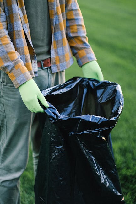 A person wearing a green and yellow plaid shirt, with a grey t-shirt underneath, is standing outdoors on a grassy area. They are holding open a large black garbage bag lined with a shiny black plastic interior. The individual is using light green gloves to grasp the edges of the bag, preparing for rubbish collection or waste disposal. The background shows a well-maintained lawn with short, green grass, suggesting a garden, park, or estate setting. The scene implies a scenario of private waste handling or on-site rubbish removal, which is an alternative to municipal collection services. The person's attire and the arrangement of the garbage bag suggest a focus on waste management or tidying up a property or outdoor space, consistent with rubbish removal activities coordinated by professional services like those offered by rubbishcollectionhammersmith.co.uk. The lighting appears natural, indicating daytime, and the environment looks clean and orderly, emphasizing a responsible approach to waste disposal and environmental care.
