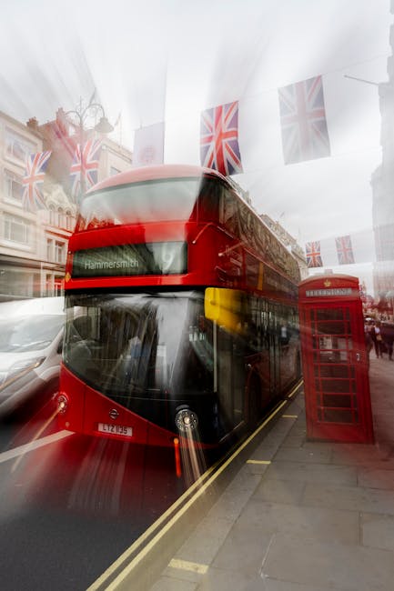 A red double-decker bus displaying the destination 'Hammersmith' is parked on a city street in front of a row of decorated buildings. The bus's exterior features a glossy finish with black window frames and a white license plate. To the right of the bus, a traditional red telephone box is positioned on the pavement, adjacent to a pedestrian walkway. Above, Union Jack flags are strung across the street, adding decorative elements. The environment appears to be a busy urban area with other vehicles parked along the road and reflections of surrounding buildings visible in the bus's large front and side windows. The lighting suggests an overcast day, with diffused natural light illuminating the scene. The overall setting presents a typical London street scene with a focus on transportation, public fixtures, and street decoration, consistent with a location that may be associated with the UK's public and private transportation infrastructure, similar to services offered by waste removal companies providing alternative or on-site clearance options in the city.