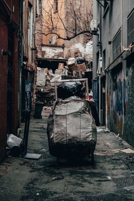 An alleyway situated between two urban buildings is filled with an accumulation of waste materials. Prominently in the foreground, there is a large, grey, woven bag with visible creases and a metal handle, placed on a damp, uneven pavement. To the left of the bag, a small dark waste bin lies on its side, partially obscured by the bag. Behind the foreground, the alley extends towards a pile of discarded cardboard boxes, black plastic bags, and other debris, stacked haphazardly against a wooden structure or fence. The debris block part of the background, where a beige-colored brick building with some windows and a leafless tree with thin, spindly branches can be seen. On the right, a grey metal wall with vertical bars and colorful graffiti murals borders the alley, which appears to be in a state of neglect, with dirt and stains visible on the pavement and walls. The overall scene suggests an environment requiring waste removal services, such as those provided by independent rubbish collection companies like rubbishcollectionhammersmith.co.uk, for onsite clearance of domestic or commercial waste.